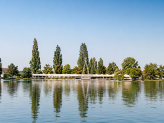 Ruhiger Badesee mit spiegelndem Wasser, grünen Bäumen und einer langen Badeanlage am Ufer bei sonnigem Wetter.