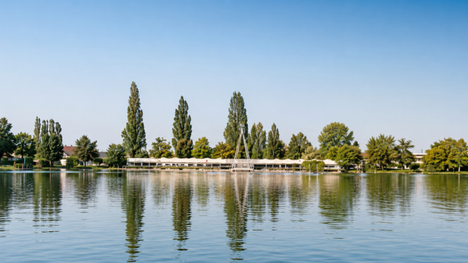 Ruhiger Badesee mit spiegelndem Wasser, grünen Bäumen und einer langen Badeanlage am Ufer bei sonnigem Wetter.