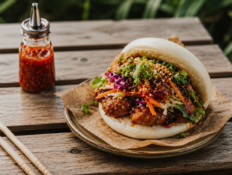 Realistisch fotografierter Bao Bun mit würziger Füllung, frischem Gemüse, Kräutern und Sesam auf einem Holztisch neben Chili-Sauce und Essstäbchen.