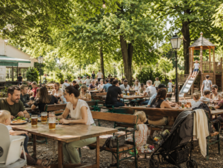 Familien sitzen in einem grünen Wiener Gastgarten, während Kinder auf einem nahe gelegenen Spielplatz spielen