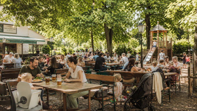 Familien sitzen in einem grünen Wiener Gastgarten, während Kinder auf einem nahe gelegenen Spielplatz spielen