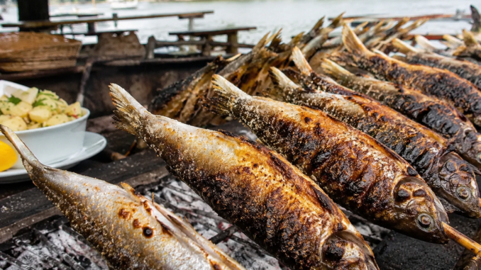 Mehrere Steckerlfische werden über Holzkohle gegrillt, im Hintergrund ist ein Gewässer zu sehen