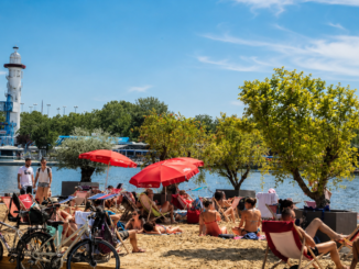 Sommerlicher Stadtstrand an der Alten Donau in Wien mit Liegestühlen, roten Sonnenschirmen, Badegästen und Leuchtturm im Hintergrund.