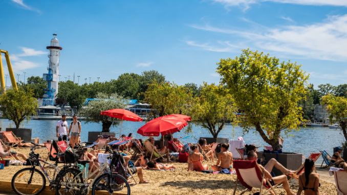 Sommerlicher Stadtstrand an der Alten Donau in Wien mit Liegestühlen, roten Sonnenschirmen, Badegästen und Leuchtturm im Hintergrund.