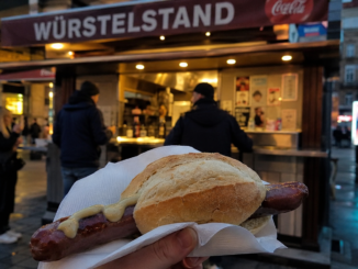 Käsekrainer im Brot mit Senf vor einem beleuchteten Wiener Würstelstand am Abend